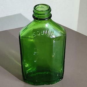 Peut inclure: Une bouteille en verre vert vintage avec un corps rectangulaire et un col court. Le mot "SQUIBB" est gravé sur le devant. La bouteille est vide et repose sur une surface de ton neutre.