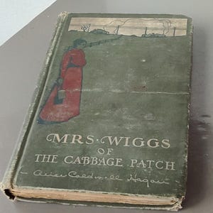 Puede incluir: Libro antiguo titulado "Mrs. Wiggs of the Cabbage Patch" con una cubierta verde. La portada presenta una ilustración de una persona con un vestido rojo y un paisaje. El título está en letras vintage elegantes.