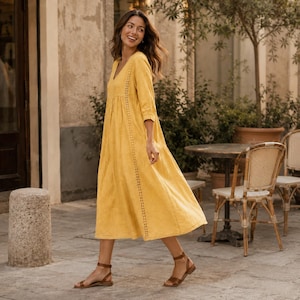 May include: A long, mustard yellow dress with three-quarter length sleeves, a V-neckline, and vertical lace-like trim detailing running down the front. The dress features a loose, flowing skirt and is paired with simple brown strappy sandals.