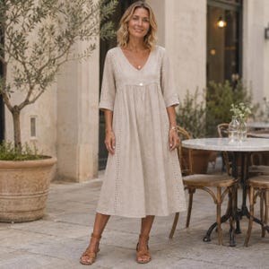 May include: A beige midi-length dress with a V-neckline, three-quarter length sleeves, and vertical lace trim detailing along the bodice. The dress features a loose, flowing silhouette and a single button closure at the center of the chest.