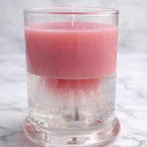 May include: A pink candle in a clear glass. The candle is a solid pink color on top and appears to be suspended in a clear liquid below. A white wick extends from the top of the candle. The glass sits on a clear, rounded base.