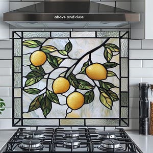 May include: Stained glass panel featuring a lemon branch with yellow lemons and green leaves. The artwork is framed and set against a white background. The panel is installed above a black stove in a kitchen setting.