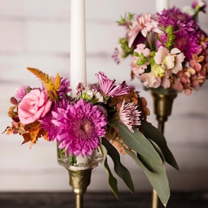 May include: Two gold candle holders with white candles and floral arrangements. The flowers are pink, purple, and green.