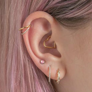 May include: An ear featuring several pieces of jewellery. A gold heart-shaped earring is in the centre, with a small pink stud below. A double-banded gold earring with small stones sits at the top, and a small hoop earring with stones is at the bottom.