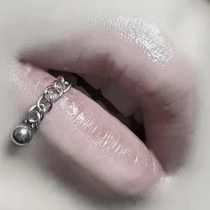 May include: Close-up of lips with a silver chain lip ring. The lip ring has a small ball at the end of the chain. The lips are glossy and a pale pink colour.