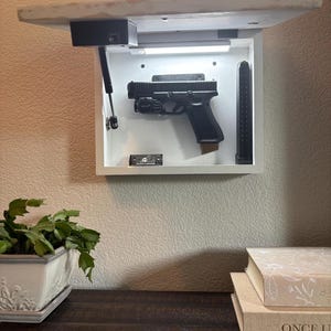May include: A wall-mounted, white gun safe with a black handgun and magazine inside. The safe has a built-in light and is mounted under a shelf. The safe is open, revealing the firearm and accessories.