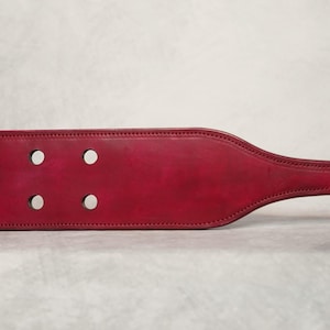 May include: A red leather paddle with eight circular holes and a handle. The paddle is a deep red color with visible stitching around the edges. A small tag is attached to the handle.