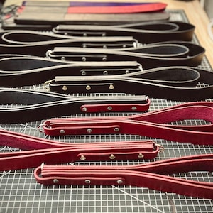 May include: An assortment of leather products, including black and red leather straps and paddles. The items are arranged on a grid surface, showcasing the craftsmanship and variety of designs. The paddles have a wooden handle.