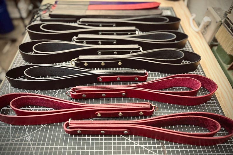 Pode incluir: Uma variedade de artigos de couro, incluindo correias e p&aacute;s de couro preto e vermelho. Os artigos est&atilde;o dispostos numa superf&iacute;cie de grelha, mostrando o artesanato e a variedade de designs. As p&aacute;s t&ecirc;m um cabo de madeira.