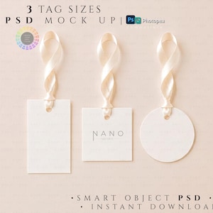 May include: Three white paper tags in rectangular, square, and circular shapes, each featuring a cream-colored satin ribbon loop at the top. The square tag displays the text NANO in a thin, dark font. The image includes the text 3 TAG SIZES PSD MOCK UP.