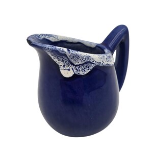 May include: A small, navy blue ceramic pitcher with a white, textured rim. The pitcher has a curved handle and a rounded body. The white rim has a bubbly, foamy appearance. The pitcher is likely used for serving liquids.