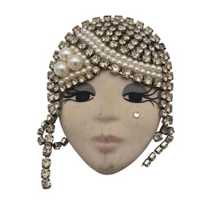 May include: Vintage face-shaped brooch with a flapper-style design. The brooch features a cream-colored face with painted eyes and lips, adorned with rhinestones, pearls, and a rhinestone-studded headpiece. A single rhinestone accents the cheek.