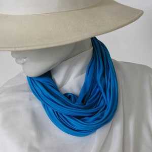 Può includere: Un cappello bianco a tesa larga e una sciarpa infinita blu brillante. La sciarpa è realizzata in un tessuto morbido ed elastico e viene indossata intorno al collo.