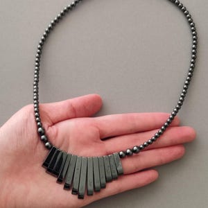 May include: A dark grey necklace featuring a beaded chain and a fan-shaped pendant. The pendant is composed of rectangular, polished stones. The necklace is shown on a hand against a grey backdrop. Jewellery.