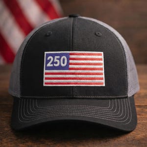 May include: A black trucker hat with a gray mesh back and a curved brim. The front of the hat features a patch with the number "250" over a red and white striped design, resembling a flag.