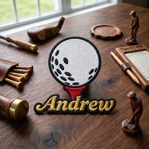 May include: A golf-themed patch with the name "Andrew" embroidered in gold and black. The patch features a white golf ball with black dimples on a red tee. Wooden golf accessories, including a score card and golf clubs, surround the patch.