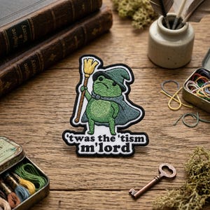 May include: Embroidered patch featuring a green frog dressed as a wizard, holding a broom. The frog wears a hat and cape, with the text "'twas the 'tism m'lord" below. The patch has a white border.