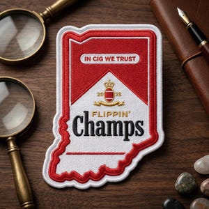 May include: Embroidered patch in the shape of Indiana, featuring a red and white design reminiscent of a cigarette pack. Text includes "IN CIG WE TRUST" and "FLIPPIN' Champs". A crown and the numbers "20 25" are also present.
