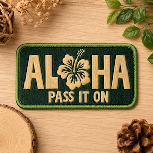 May include: A rectangular embroidered patch with a dark green background and a thick green border. The text ALOHA is stitched in tan thread, with a tan hibiscus flower replacing the letter O. Below, the text PASS IT ON is stitched in tan.