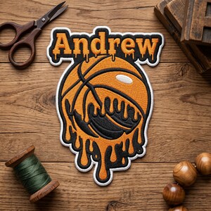 May include: Embroidered patch featuring a basketball with a melting design in orange and black, with the name "Andrew" above. The patch has a white border. The image is on a wooden surface with scissors and a spool of thread.