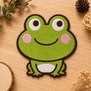 May include: An embroidered iron-on patch shaped like a frog face. The frog is bright green with a dark brown border, large white eyes with black pupils, pink circular cheeks, a curved black mouth, and a white oval patch on its belly.