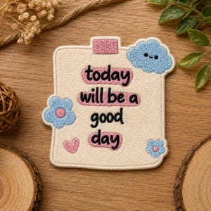 May include: A rectangular embroidered patch with a cream-colored background featuring the black text today will be a good day. The patch includes pink rectangular accents, a blue cloud with a face, two blue flowers with pink centers, and a pink heart.