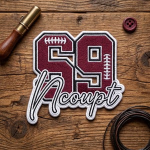 May include: A maroon and white embroidered patch with the number "59" in a football design, with the word "Accept" in script. The patch is on a wooden surface, with a button and leather cord nearby.