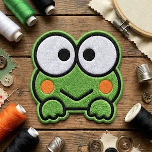 May include: Embroidered green frog patch with large white eyes, black outlines, and orange cheek spots. The patch is surrounded by sewing supplies, including spools of thread, buttons, and a wooden embroidery hoop, on a wooden surface.