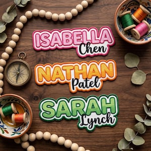 May include: Three embroidered name patches: "Isabella Chen" in pink, "Nathan Patel" in orange, and "Sarah Lynch" in green. The patches are on a wooden surface, along with a compass, spools of thread, and wooden beads.