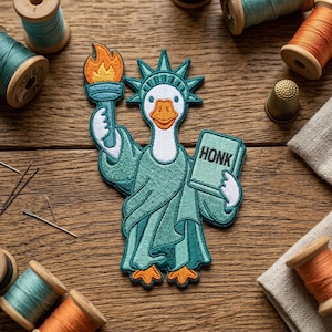 May include: Embroidered patch featuring a goose dressed as the Statue of Liberty, holding a torch and a book that says "HONK". The goose is wearing a green robe and crown. The patch is surrounded by spools of thread.
