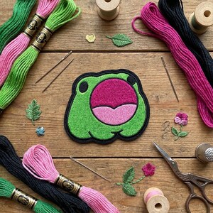 May include: A green frog patch with a pink mouth and black outline is surrounded by embroidery supplies. Pink, green, and black embroidery floss, needles, scissors, and a thimble are arranged on a wooden surface, suggesting a crafting theme.