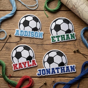 May include: Four embroidered soccer ball patches with names: Addison, Ethan, Kayla, and Jonathan. Each patch features a black and white soccer ball design above a name in a different color: blue, green, red, and blue. The patches are on a wooden surface.