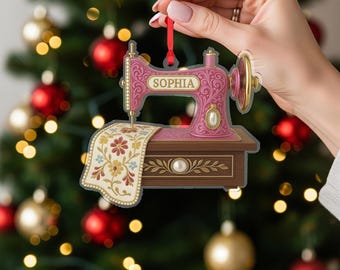May include: A pink sewing machine Christmas ornament with the name "SOPHIA" on it. The ornament has a floral design on the machine and a decorative fabric piece. The ornament is held by a hand in front of a Christmas tree.