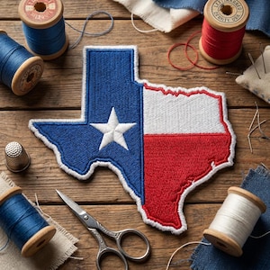 May include: Embroidered patch in the shape of Texas, featuring the state flag design. The patch is blue with a white star and red and white stripes. Sewing supplies, including spools of thread, scissors, and a thimble, surround the patch.