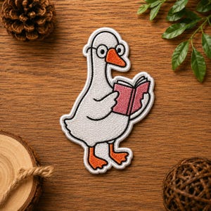 May include: An embroidered patch featuring a white goose with an orange beak and feet, wearing round black glasses and holding an open pink book. The design is outlined in white thread, showcasing detailed stitching on the bird and the book.
