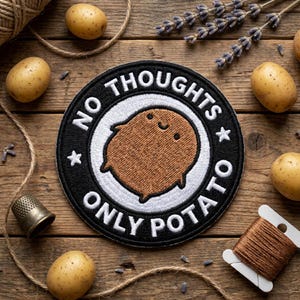 May include: A round, black and white patch with the text "NO THOUGHTS ONLY POTATO" encircling a smiling cartoon potato. The patch is surrounded by potatoes, twine, and sewing notions on a wooden surface.
