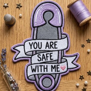 May include: Embroidered patch in the shape of a safety pin, with the words "YOU ARE SAFE WITH ME" on a banner. The patch is primarily purple, gray, and white, with a small pink heart. Decorative elements include a thimble, thread spool, and stars.