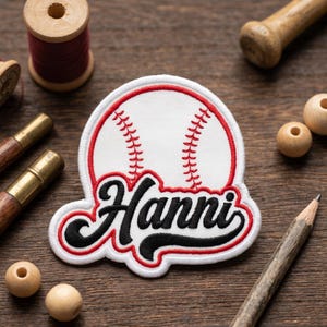 May include: A white baseball patch with red stitching and a red border. The patch features the name "Hanni" in black script with a red outline. The patch is surrounded by sewing supplies on a wooden surface.