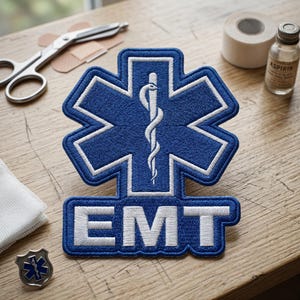 May include: A blue and white embroidered EMT patch featuring the Star of Life symbol with a staff and serpent. The word "EMT" is embroidered in white below the symbol. Other medical supplies, including scissors, tape, and a small bottle of aspirin, are also visible.
