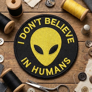 May include: A black circular patch with yellow embroidered text that reads "I DON'T BELIEVE IN HUMANS" around a yellow alien head. The patch is surrounded by sewing supplies, including spools of thread, buttons, and scissors.