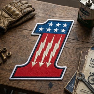 May include: Embroidered patch in the shape of the number one, featuring a blue top section with white stars and a red lower section with white lightning bolts. The patch has a white border and is on a wooden surface.