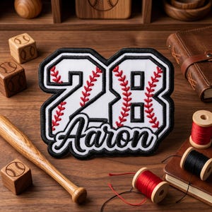 May include: A white baseball patch with the number 28 and the name "Aaron" in black script. The numbers are outlined in black with red stitching, resembling baseball stitching. The patch is on a wooden surface with baseball-themed accessories.
