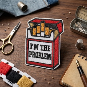 May include: Embroidered patch in the shape of a cigarette pack, with the text "I'M THE PROBLEM" in white. The pack is red with a white border and contains several yellow cigarettes. The patch is on a wooden surface with sewing tools.
