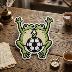 May include: Embroidered patch featuring two green frogs with raised arms, holding a black and white soccer ball. The patch has a white border. The frogs have light green bodies and dark green outlines. A fun, whimsical design.