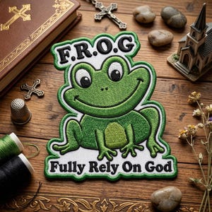 May include: Embroidered patch featuring a smiling green frog with the text "F.R.O.G. Fully Rely On God" in black and white. The patch has a white border and is set against a wooden background with religious items.