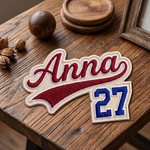 May include: A custom embroidered patch with the name "Anna" in a script font, with a red and white color scheme. Below the name is a red banner. A separate patch with the number "27" in blue is also visible.