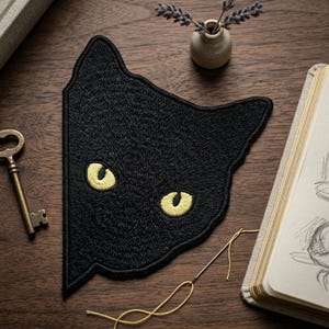 May include: A black cat face patch with yellow eyes. The patch is embroidered and has a detailed design. The image also includes a key, a small vase with lavender, a notebook, and a needle with yellow thread.
