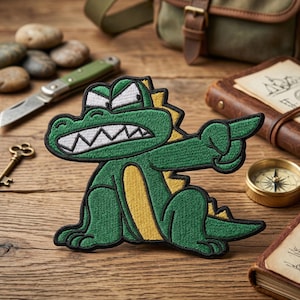 May include: Embroidered patch featuring a green alligator with a yellow belly and accents, pointing with its right arm. The alligator has a white-toothed grin and white angry eyebrows. The patch is on a wooden surface.
