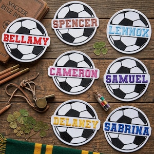 May include: Circular soccer ball patches with personalized names embroidered in various colors. Names include Bellamy, Spencer, Lennox, Cameron, Samuel, Delaney, and Sabrina. The patches are on a wooden surface with soccer-related accessories.