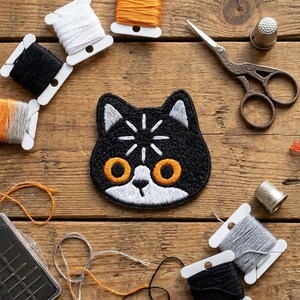 May include: A black cat-shaped embroidered patch with orange eyes and white accents. The patch is surrounded by spools of thread in various colors, scissors, thimbles, and a needle case, all arranged on a wooden surface. A craft-themed item.